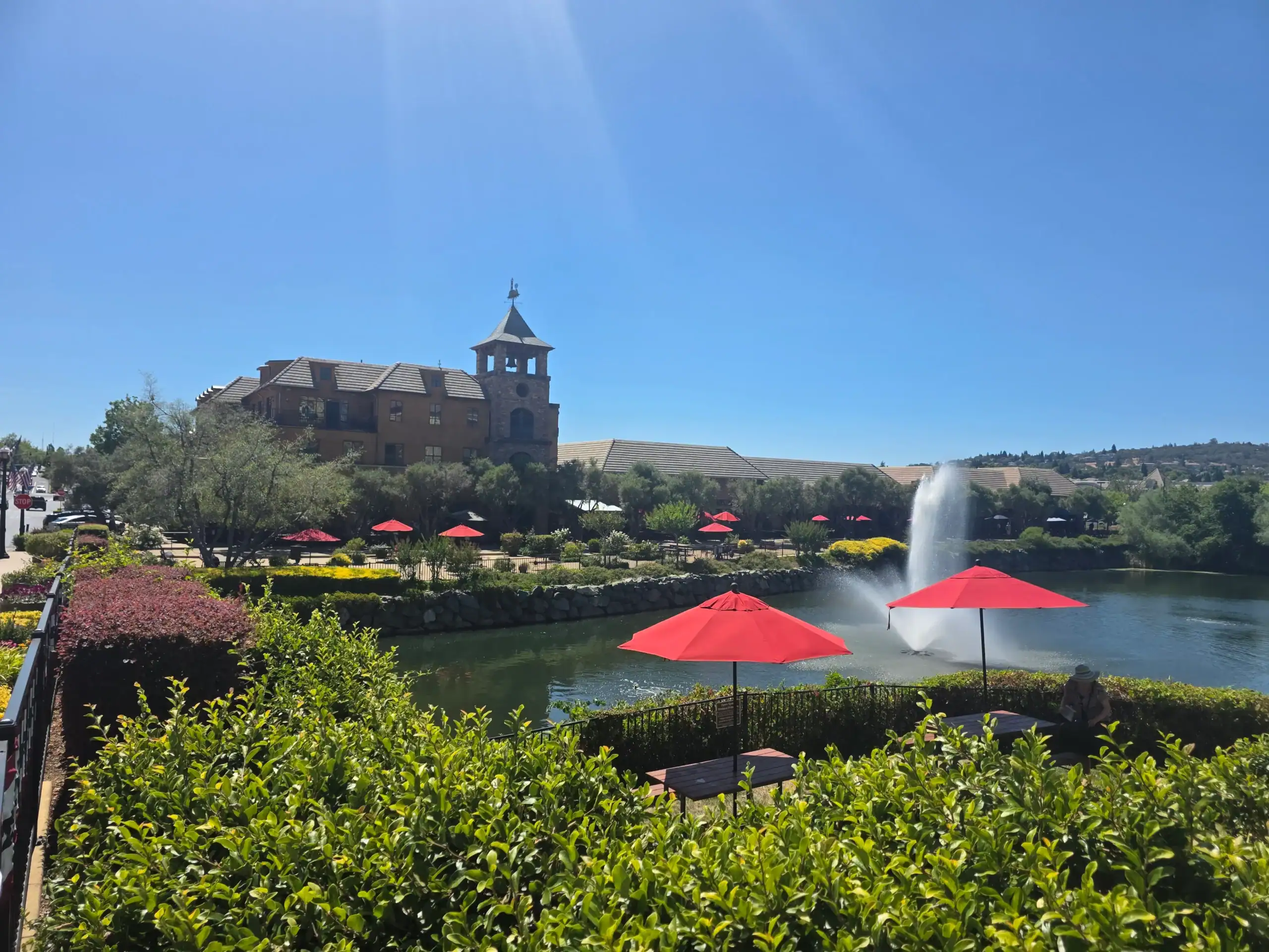 Panaramic view of pond and fountain in El Dorado Hills Town Center Scenic view of a park with a pond, red umbrellas, lush greenery, and a historic building with a tower under a clear blue sky.